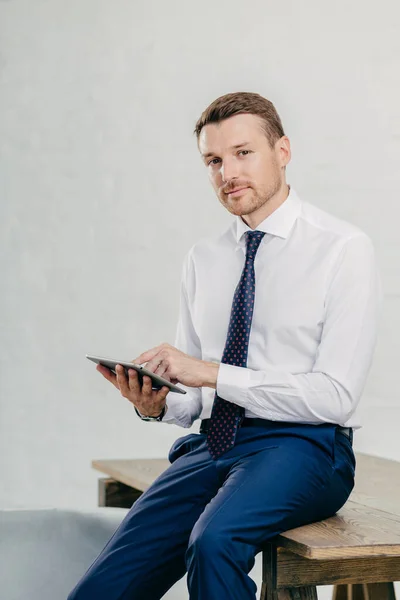 Serious male entrepreneur watches video on digital tablet, messages with business partners, dressed elegantly, sits on wooden table, poses against white wall. Businessman updates banking account