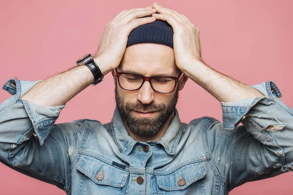 Isolated shot of unhappy desperate man with stubble keeps hands on head, wears fashionable clothing: denim jacket, hat and eyewear, poses against pink background. Forgetful male poses indoor