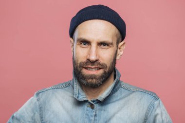 Kalın sakal ve bıyık memnun erkek model fotoğraf makinesi içine emin ifade ile görünüyor, şık şapka ve pembe arka plan üzerinde izole ceket giyer. İnsanlar, duygular, yüz ifadeleri