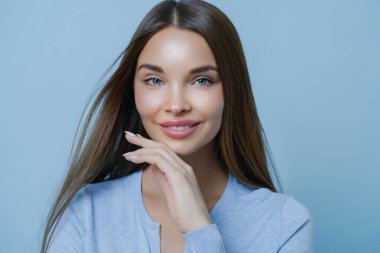 Kaygısız Avrupalı kadın model çene hattına dokunuyor, dişleriyle gülümsüyor, sağlıklı cildine sahip, makyajı az, rüya gibi bakışları var, ilginç bir şeyle yakalanmış, mavi arkaplan üzerinde izole edilmiş.