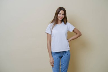 Çekici genç kadın elini belinde tutuyor, günlük giysiler giyiyor, hafta sonu dışarı çıkmaya hazır, güzel havadan hoşlanıyor, birbirine olumlu bir tavır sergiliyor, bej duvarın üzerinde poz veriyor.