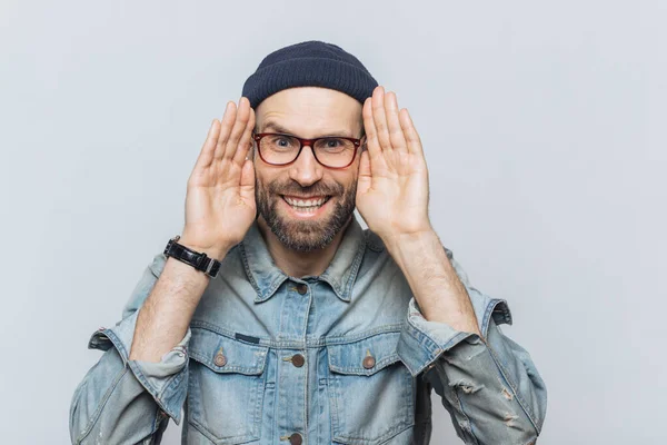Portrait of happy hipster guy with thick beard and mustache wears stylish hat and glasses, keeps hands near face, tries to see something in darkness, isolated over grey background, has fun indoor