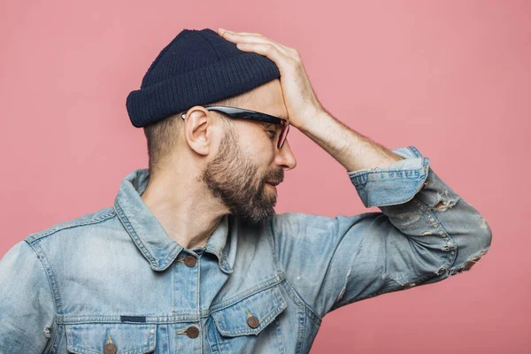 Sideways portrait of bearded male has regretful expression, keeps hand on forehead, looks desperately, isolated over pink background. Stressful unshaven man in glasses has terrible headache.