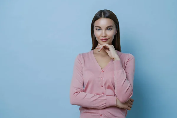 Waist up shot of beautiful young European woman keeps hand under chin, looks directly at camera, applies makeup, listens interlocutor carefully, wears casual jumper, isolated on blue background.