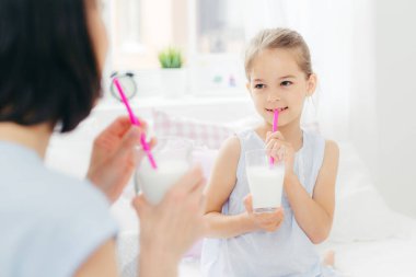 Back view of young brunette mother looks at her small daughter, drink fresh milk cocktails in bed, have delighted expressions and pleasant talk during morning, Parenthood and leisure concept
