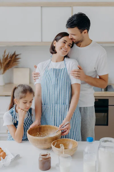 Family cook meal at home. Husband and wife embrace with love, small girl poses near, prepare delicious dinner, use milk, chocolate, eggs, make homemade pastry, pose against kitchen interior.
