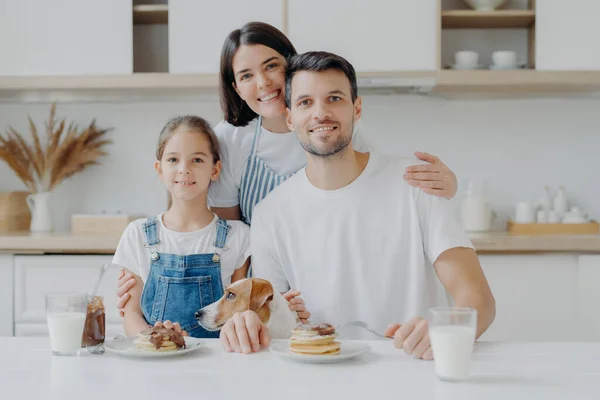 Happy family and dog pose in cozy kitchen, eat fresh homemade pancakes with chocolate and milk, look positively at camera. Mother in apron embraces husband and daughter, likes cooking for them