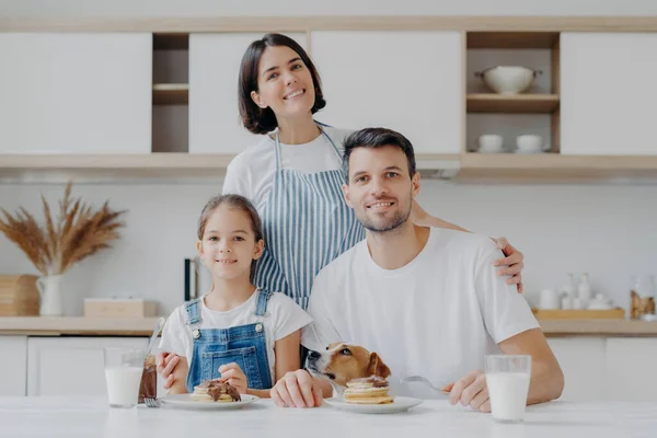 Three family member and their pet pose all together at kitchen, eat sweet pancakes with chocolate, drink milk, affectionate housewife poses behind husband and daughter, embraces, prepares breakfast