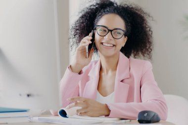 Güzel kıvırcık saçlı, esmer bir kadın iş arkadaşıyla telefonda konuşmaktan hoşlanıyor, mutlu bir yüz ifadesi var, masaüstünde oturuyor, gözlük takıyor ve pembe takım elbise giyiyor, sıkı çalışmadan sonra mola veriyor. Teknoloji