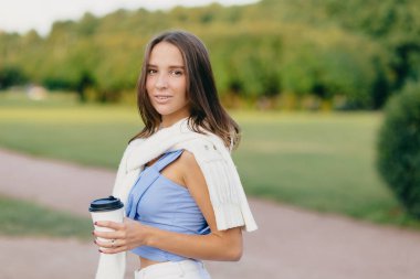Horizontal shot of brunette lady with slim body, dressed in t shirt, white sweater on shoulders, drinks aromatic coffee, stands outdoor against green nature background. People and lifestyle concept