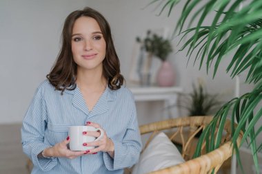 Koyu saçlı, sağlıklı yumuşak deri, memnun çekici kadın kapalı atış sabah kahve içer, pijama giyiyor, düşünceli bir yana görünüyor, rahat oda pozlar, rahat hissediyor. Boş zaman kavramı