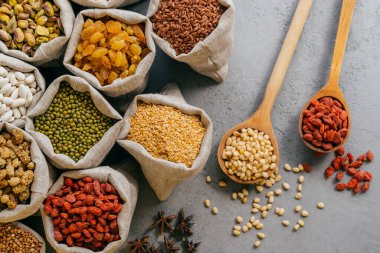 Çeşitli kurutulmuş meyve ve tahıllar paketlenmiş küçük çuvallar. Gri arka planda iki tahta kaşık. Sağlıklı baklagiller ve tahıllar