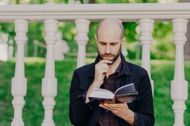 Odaklı ciddi sakallı kel erkek çene tutar, boş zaman ve güneşli yaz günü açık kitap, beyaz balkon, arsa üzerinde konsantre karşı açık pozlar sahiptir dikkatle okur. Park'ta dinlenme.