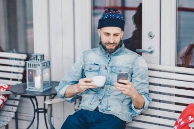 İnsanlar, eğlence ve teknoloji kavramı. Pozitif sakallı genç erkek recreats teras kafede tutan modern akıllı telefon, ileti online ile sokulgan, içecek çay veya kahve, denim ceket giyer