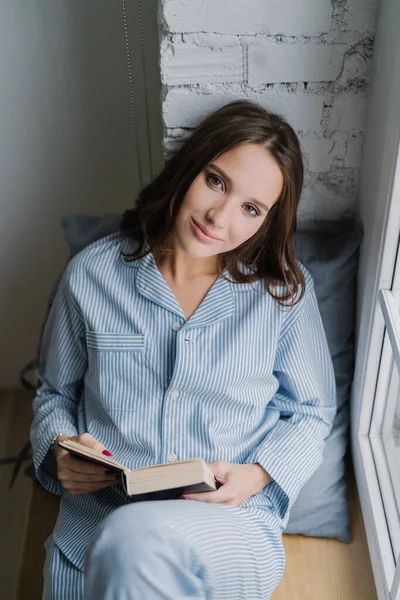 Relaxed young of beautiful in casual clothes, sits at window near brick wall, back to soft pillow, reads interesting detective story, looks directly at camera, wears casual costume. People and reading