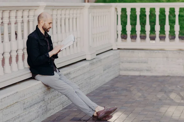 Concentrated bearded middle aged male reads book with great interest, finds out new interesting facts, leans at white balcony, enjoys calm atmosphere and his hobby. People and leisure concept
