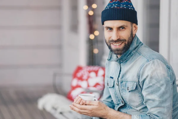 Portrait of handsome young smiling male enjoys spare time, holds mug of coffee, being deep in thoughts, has coffee break during shining warm day, focused on something. Pleased bearded stylish man