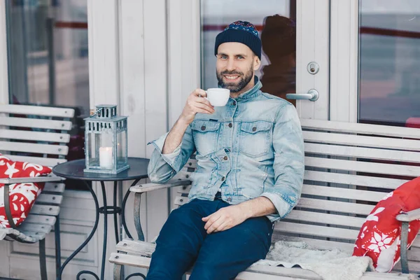 Happy blue eyed male with positive look, sits against cozy interior at terrace cafeteria, enjoys aromatic coffee, has spare time after work. Handsome man has beard, looks joyfully at camera.