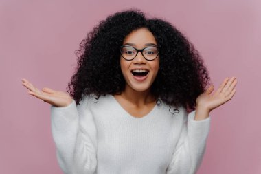Neşeli kıvırcık bayanın portresi kollarını kaldırır ve avuçlarını açar, optik gözlük takar ve beyaz kazak giyer, yakın kişiden hoş bir sürpriz alır, menekşe stüdyo duvarında izole olur. Mutluluk