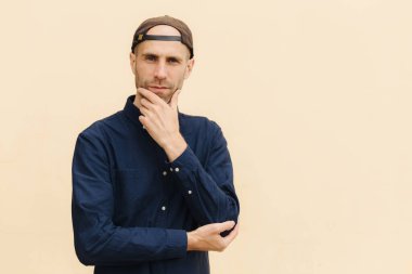 Confident European male holds chin, dressed in shirt and cap, has serious expression, poses against beige background. Handsome man thinks about something. People, lifestyle, masculinity concept