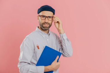 Şık uzak çalışan şeffaf gözlük, şık şapka, gömlek giyer, mavi ders kitabı tutan, bilgi yazmak için kalem kullanır, pembe arka plan üzerinde teşkil etmektedir. Memnun erkek yazar bilgileri analiz eder.