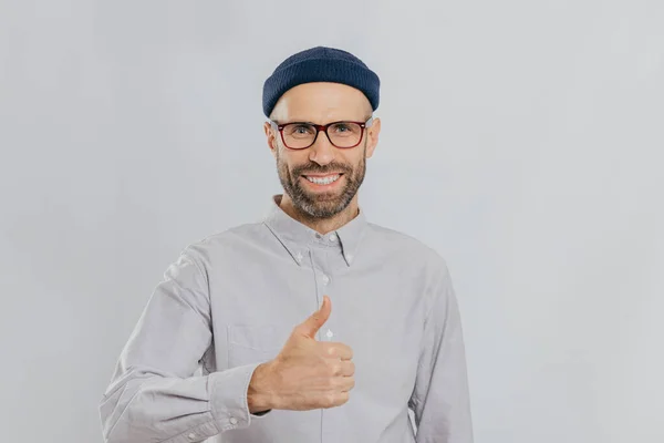 Positive smiling man with stubble, raises thumb up, demonstrates his like and approvement, wears headgear and formal shirt, isolated over white background. My answer yes. Gesturing. Body language
