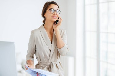 Mutlu, düşünceli, başarılı kadın çalışan, modern cep telefonunu kulağının dibinde tutuyor, telefon görüşmesi yapıyor, kenara bakıyor, zarif bir şekilde giyiniyor, aylık raporları gözden geçiriyor, grafikleri çalışıyor, ofiste duruyor.