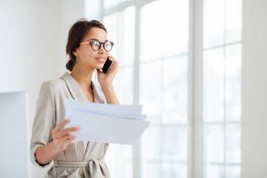 Düşünceli iş kadını kameraya yan bakıyor, kağıt belgeler tutuyor, telefonla konuşuyor, mali rapor hazırlıyor, birlikte çalışma alanında poz veriyor, optik gözlük takıyor ve resmi takım elbise giyiyor. Belgeler..