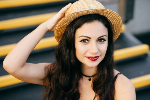 Beautiful woman with dark eyes and hair having nice make-up wearing straw hat sitting at stairs having confident look. Attractive brunette female with appealing appearance posing into camera