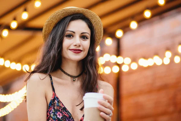 stock image Portrait of brunette lady with charming eyes, well-shaped red lips and pure skin, dressed in summer hat and shirt, holding fresh takeaway coffee in paper cup, relaxing at terrace of cozy cafe