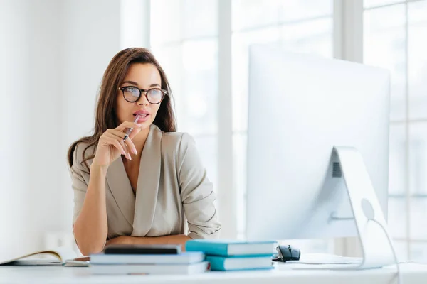 沉默寡言的女企业家把笔收在嘴里 专注于计算机监控 思考新战略的发展 穿着雅致的服装和眼镜 面对办公室的内部摆设 — 图库照片