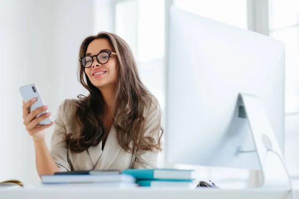 穿着企业服装微笑的女性通过智能手机观看滑稽视频 坐在办公室的桌面上 用电脑工作 享受工作过程 浏览项目信息 戴眼镜 — 图库照片