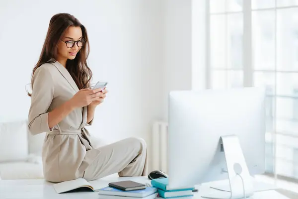 穿着米色正装的优雅女人专注地阅读网站上的新闻 独自坐在橱柜的桌旁 身边的电脑显示器和笔记本 女员工在办公室里使用现代智能手机 — 图库照片