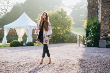 Hoş görünümlü ince uzun boylu kadın portresi giyer cape ve yüksek topuklu ayakkabı, geçti standları bacaklar, açık havada karşı antik alan teşkil etmektedir, doğrudan içine kamera kendinden emin bir görünüme sahiptir. Yaşam tarzı kavramı