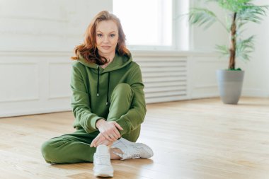Spor adamış spor kırmızı saçlı kadının yatay çekimi, yeşil eşofman ve beyaz spor ayakkabı giyer, geniş odada yerde poz lar, pilates egzersizleri, yoga pratiği sonrası dinlenir.