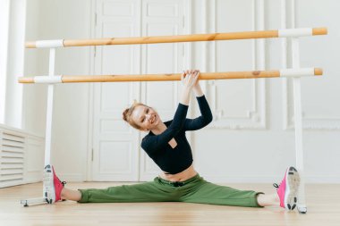 Beyaz kadın balerin bacaklarını yana açıyor, bacaklarını açıyor, barlara el ele tutuşuyor, bale sınıfına giriyor, spor kıyafetleri ve spor ayakkabıları giyiyor, mükemmel bir vücudu var, başını sağa eğiyor, kendini esnetiyor..