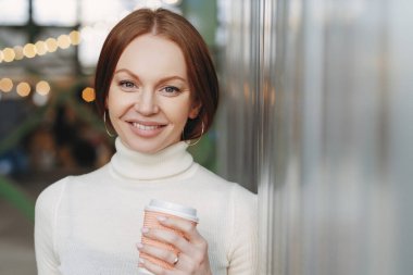 Genç erkek casual Beyaz boğazlı Kazak giymiş yakışıklı, kağıt bardak kahve ve aromatik cappucino tutar, kameraya mutlu görünüyor, dışında poz veriyor, boş zaman vardır. Yaşam tarzı kavramı