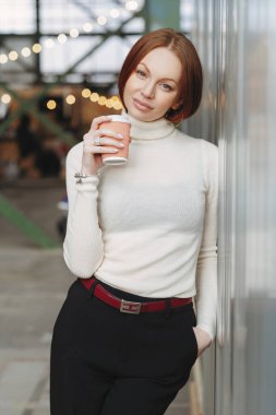 Dikey görüntü düşünceli genç kadının beyaz jumper ve siyah pantolon giyer, paket servisi olan restoran kahve tutar, bir şey hoş pozlar açık hakkında rüyalar, Eli cebinde tutuyor. İnsanlar ve yaşam tarzı