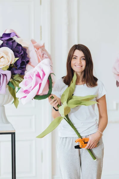 Florist at work: beautiful woman makes bouquet, works at flower shop, arranges decorations, dressed in casual outfit poses indoor. Vertical shot. Female collects roses in bunch. Small business concept