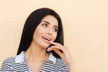 Memnun güzel genç kadın siyah saçlı mutlu görünüyor ve sevinçle bir yana, kayıtsızca giyinmiş hafif arka planı teşkil etmektedir, temiz cilt vardır, manikür, el ağız yakın tutar, studio modelleri