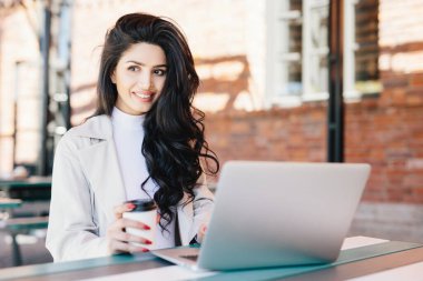 Koyu dalgalı saçlı bir kenara kafede temiz hava açık havada hayran paket servisi olan restoran kahve içme sınıfları sonra dinlenme ararken ifade memnun onu laptop haber okuma gülümseyen kız öğrenci