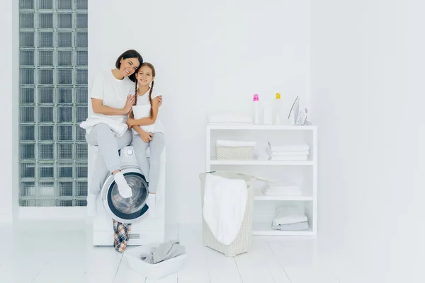 stock image Shot of beautiful woman and his small daughter embrace and smile pleasantly, sit on washing machine, wash linen in laundry room, have friendly relationship, do laundry at home. Housework concept