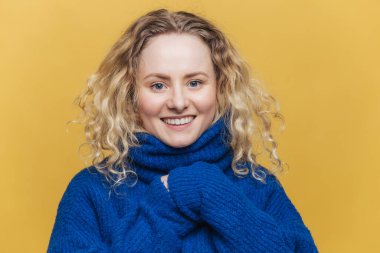 Geniş bir gülümseme, kıvırcık genç kadınla mutlu olumlu duygular ifade eder, yakalı, pozlar sarı arka plan Mavi kazak giyiyor. İyi haberler duymak iyi görünümlü kadın modeli memnun.