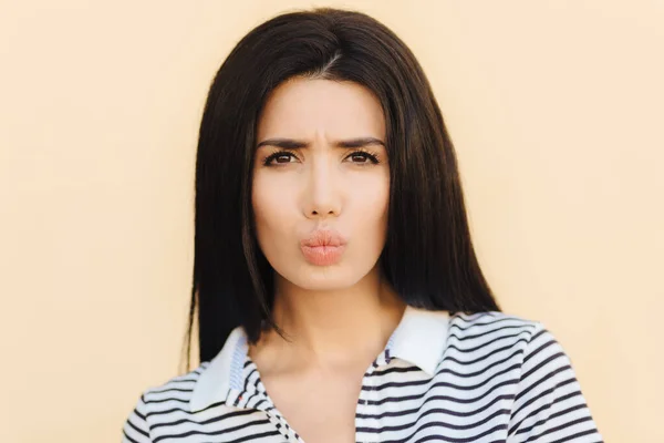 People, beauty and facial expressions concept. Discontent brunette cute woman pouts lips and frowns face, has sullen look, dressed in casual striped t shirt, has gloomy expression after quarrel