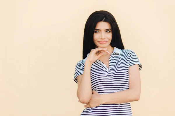Waist up portrait of thoughtful female with dark straight long hair, keeps hand under chin, contemplates about something, dressed in striped t shirt, poses against light background with blank space