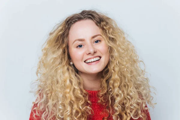 Close up shot of happy attractive woman with broad shining smile, bushy blonde curly hair, poses against white background, satisfied to pass examination session successfully. People, beauty.