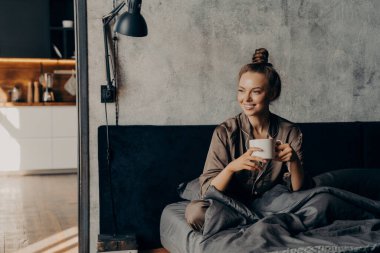 Mutlu, beyaz, pijamalı bir kadın sabah kahvesinin keyfini çıkarırken uyandıktan sonra yoğun iş gününe başlamadan önce modern bir apartman dairesinde ya da otel odasında dinleniyor.