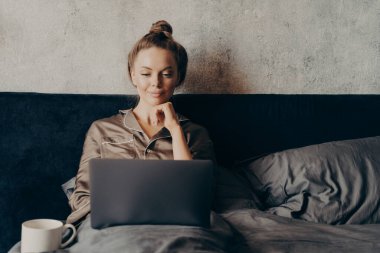 Evde tembellik ederek, evde yatıp, pijama giymiş bilgisayar monitörüne bakarken, en sevdiği filmi dizüstü bilgisayarda izlemek için internette sörf yapan pozitif ve rahat genç bir kadın.