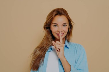 Portrait of joyful smiling female with long light brown hair holding index finger at her lips, saying 'shh', asking for silent or to keep her secret. Human face expressions, emotions and feelings