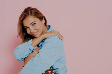 Stüdyoda, yan yana oturan, hafifçe eğilmiş başıyla gülümseyen, mavi günlük gömlek giyen, solgun pembe arka plan ve boş alan üzerine izole edilmiş genç, güzel model kadının portresini kapat.